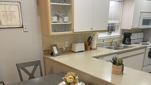 Un comedor con una mesa dispuesta para cuatro personas, rodeada de sillas de madera. Se ve una cocina al fondo con armarios blancos y una encimera.