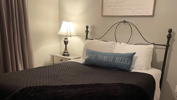 Uma cama bem arrumada com edredom preto e branco, um travesseiro azul com a inscrição 'Grateful. Thankful. Blessed.', e um quadro com uma placa na parede acima da cama.