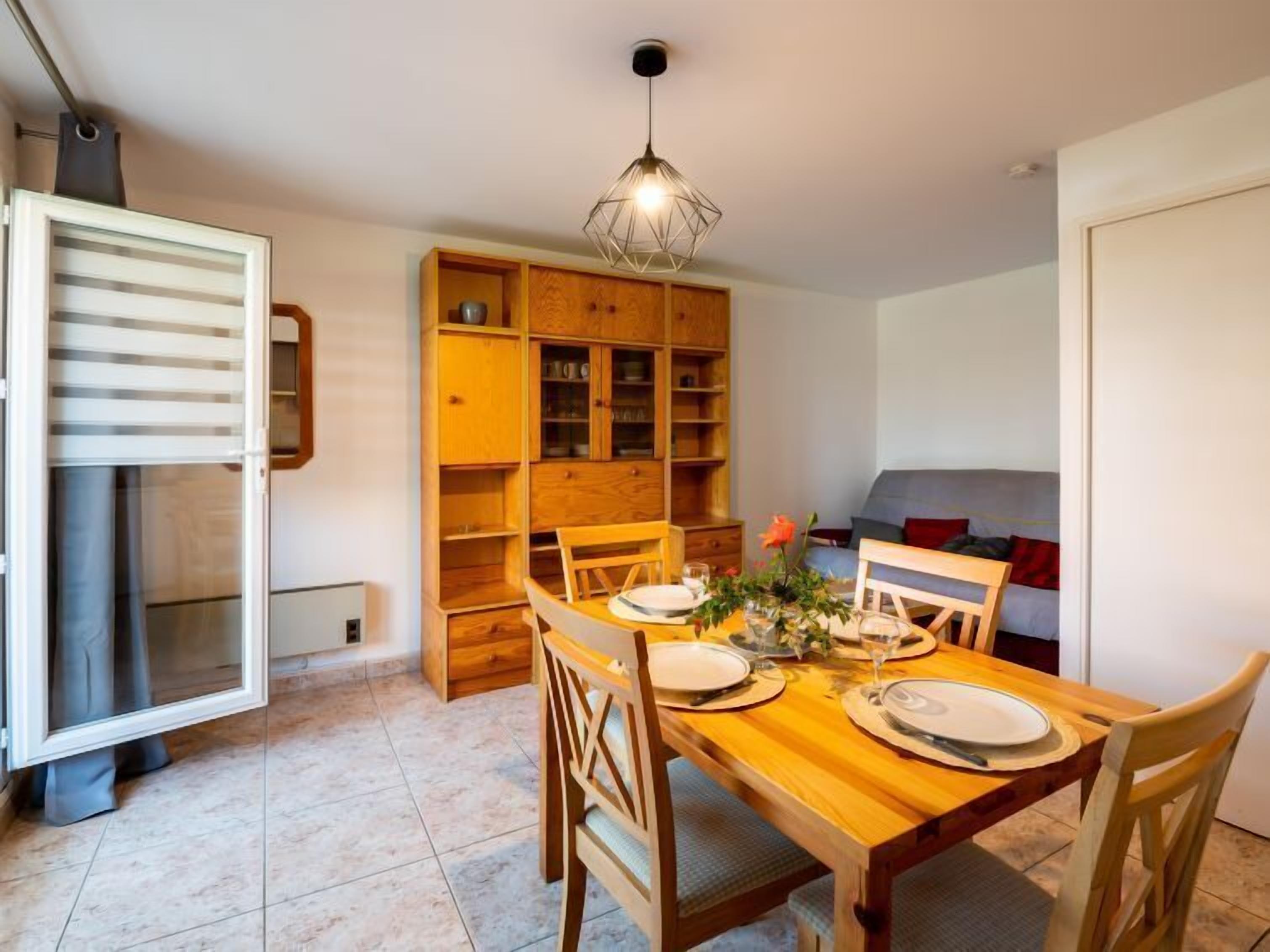 Een gezellig eetgedeelte met een houten tafel gedekt voor twee personen, omringd door bijpassende stoelen. Links staat een houten kast met open planken, en op de achtergrond is een bank zichtbaar.