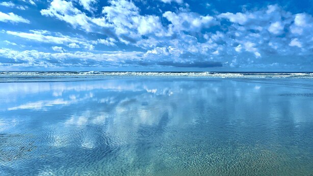 Uma praia com águas claras refletindo o céu e as nuvens.