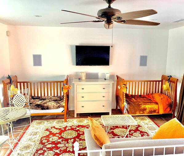 A bedroom with two cribs, a TV, a ceiling fan, a dresser, and a patterned rug.