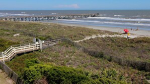 A beach with a wooden boardwalk leading to the shore, a pier extending into the ocean, and a red lifeguard chair on the sand. - Oceanfront, Top Floor/ Corner Sea Cabin, Gorgeous Views with Pool (Isle of Palms)