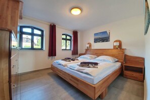 A bedroom with a wooden bed, two windows with curtains, a nightstand, and a wall-mounted lamp. - Haus Besch - Apartment Kleine Kapitänskajüte - Kleine Kapitänskajüte: 68m², 2-raum, 2 Pers., Terr., (Mönchgut)