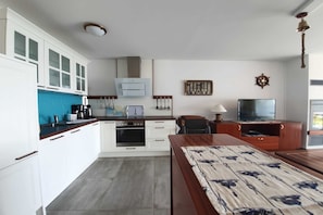 A compact kitchen with white cabinets, a stove, and a microwave. There is a dining area with a wooden table and a TV. - Haus Besch - Apartment Kleine Kapitänskajüte - Kleine Kapitänskajüte: 68m², 2-raum, 2 Pers., Terr., (Mönchgut)