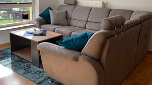 A modern living room with a sectional sofa, a small coffee table, and a view of a house through a large window. - Ferienwohnung Pusteblume - 2 Schlafzimmer, Küche, Wohnzimmer, Bad, Balkon (Edertal)