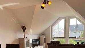 A spacious living area with a high ceiling, a large window offering a view of the neighborhood, a flat-screen TV, and a cozy seating area. - Ferienwohnung Pusteblume - 2 Schlafzimmer, Küche, Wohnzimmer, Bad, Balkon (Edertal)