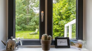 Una ventana con vista a un jardín, un cactus, una tetera, un letrero de bienvenida y una postal.