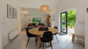 Un salón moderno con una mesa redonda de madera, sillas negras, un sofá con cojines, una lámpara colgante y vistas al jardín a través de un ventanal.