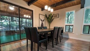 A dining area with a large table, six chairs, and a view of a forest through glass doors.