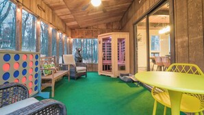 A cozy indoor space with wooden walls, a green carpet, and a ceiling fan. There are wicker chairs, a yellow table, and a play area with a bear statue.