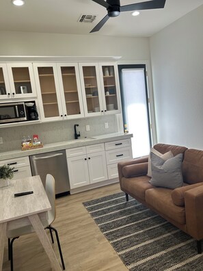 A modern kitchen with white cabinets, a built-in microwave, and a coffee maker. There's a dining table with a remote control, a brown sofa with cushions, and a striped rug.