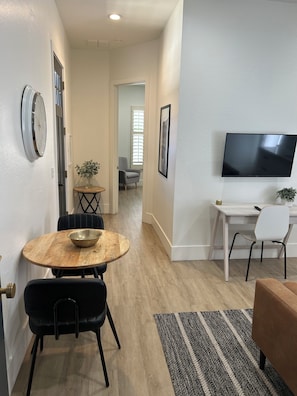 A modern living room with a round wooden table, black chairs, a TV mounted on the wall, and a patterned rug.