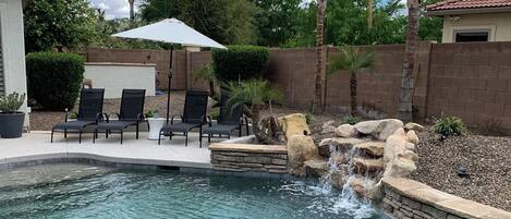 A swimming pool with a waterfall feature, surrounded by palm trees and a patio with chairs.