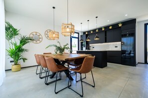 A modern kitchen with black cabinets, a wooden dining table, and brown chairs. There are hanging wicker lights and potted plants.