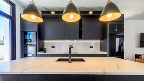 Une cuisine moderne dotée d’un grand îlot central, de meubles de rangement aux couleurs sombres et d’un éclairage suspendu.