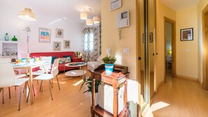 A bright living room with a red sofa, white chairs, a wooden coffee table, and a dining area with a white table and chairs. There is a potted plant on a wooden side table, and a hallway with a door leading to another room. - Beautiful & Bright Apartment for 4 People, Parking Optional (Sevilla)