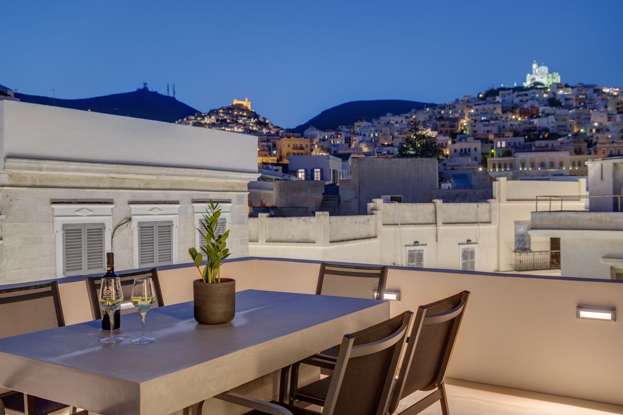 A rooftop terrace with a table set for two, overlooking a cityscape with buildings and a hill.