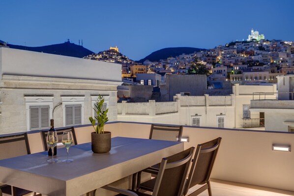 A rooftop terrace with a table set for two, overlooking a cityscape with buildings and a hill.