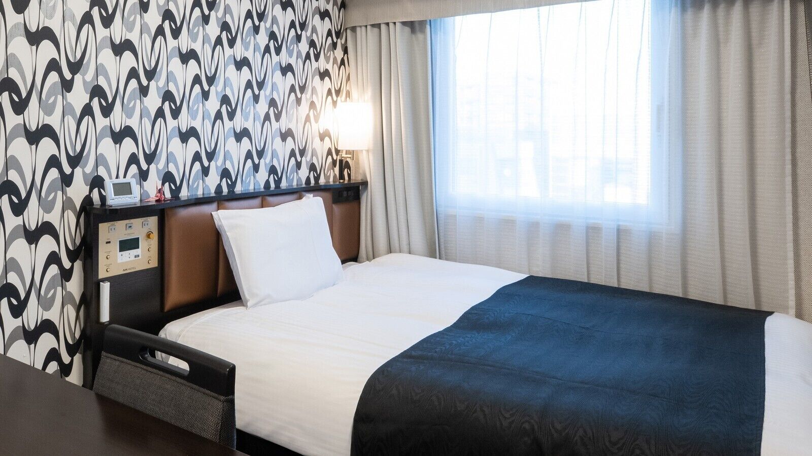 A hotel room with a bed, a control panel, a nightstand, and a window with curtains.
