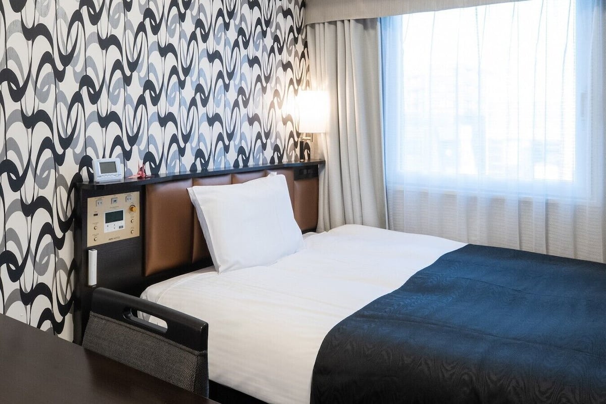 Une chambre d’hôtel avec un lit, un panneau de commande, une table de chevet et une fenêtre avec des rideaux.