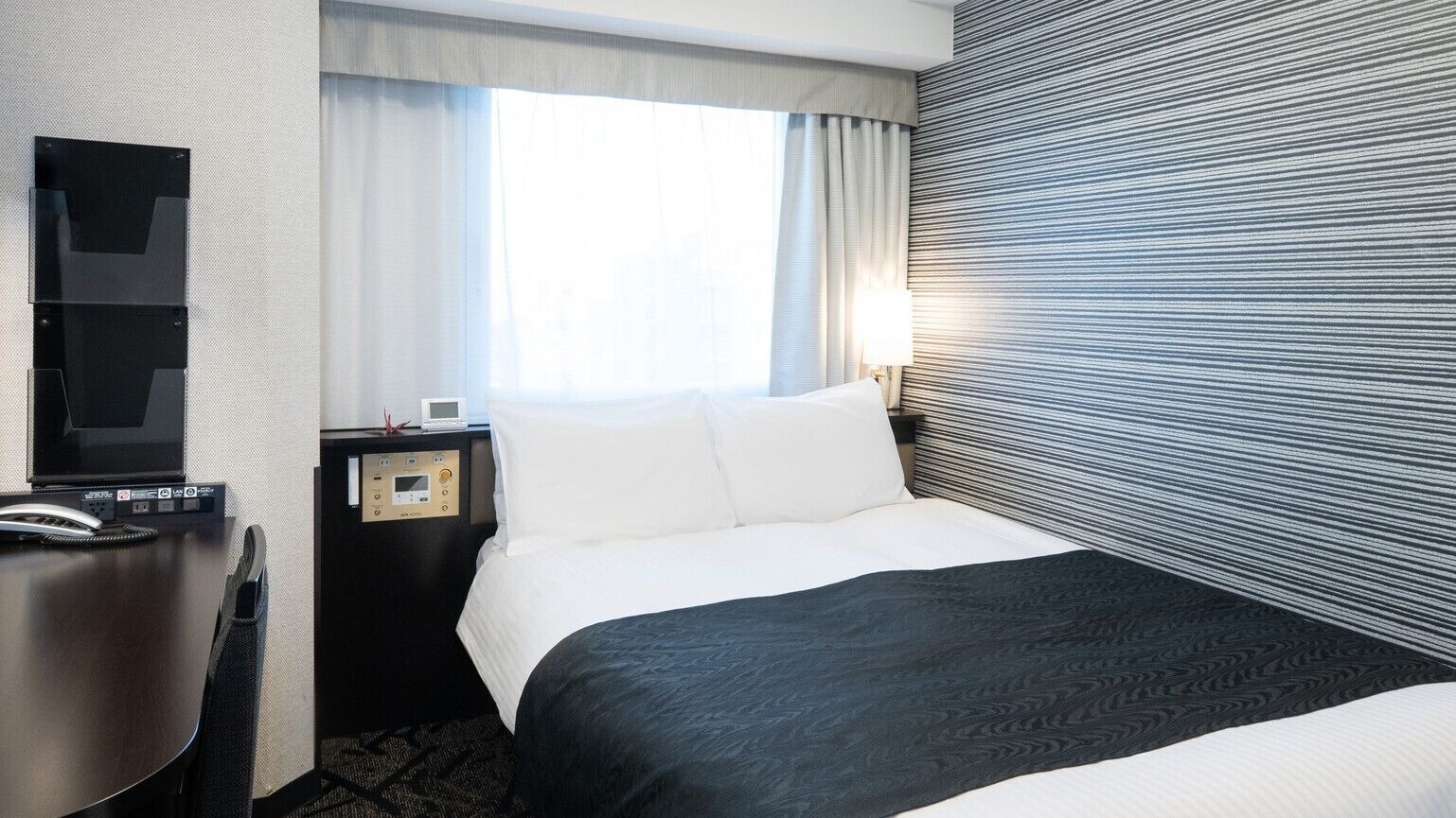 A hotel room with a bed, a desk with a telephone, a nightstand with a lamp, and a window with curtains.
