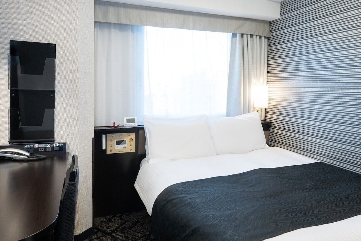 Une chambre d’hôtel comprenant un lit, un bureau avec un téléphone, une table de chevet avec une lampe et une fenêtre avec des rideaux.