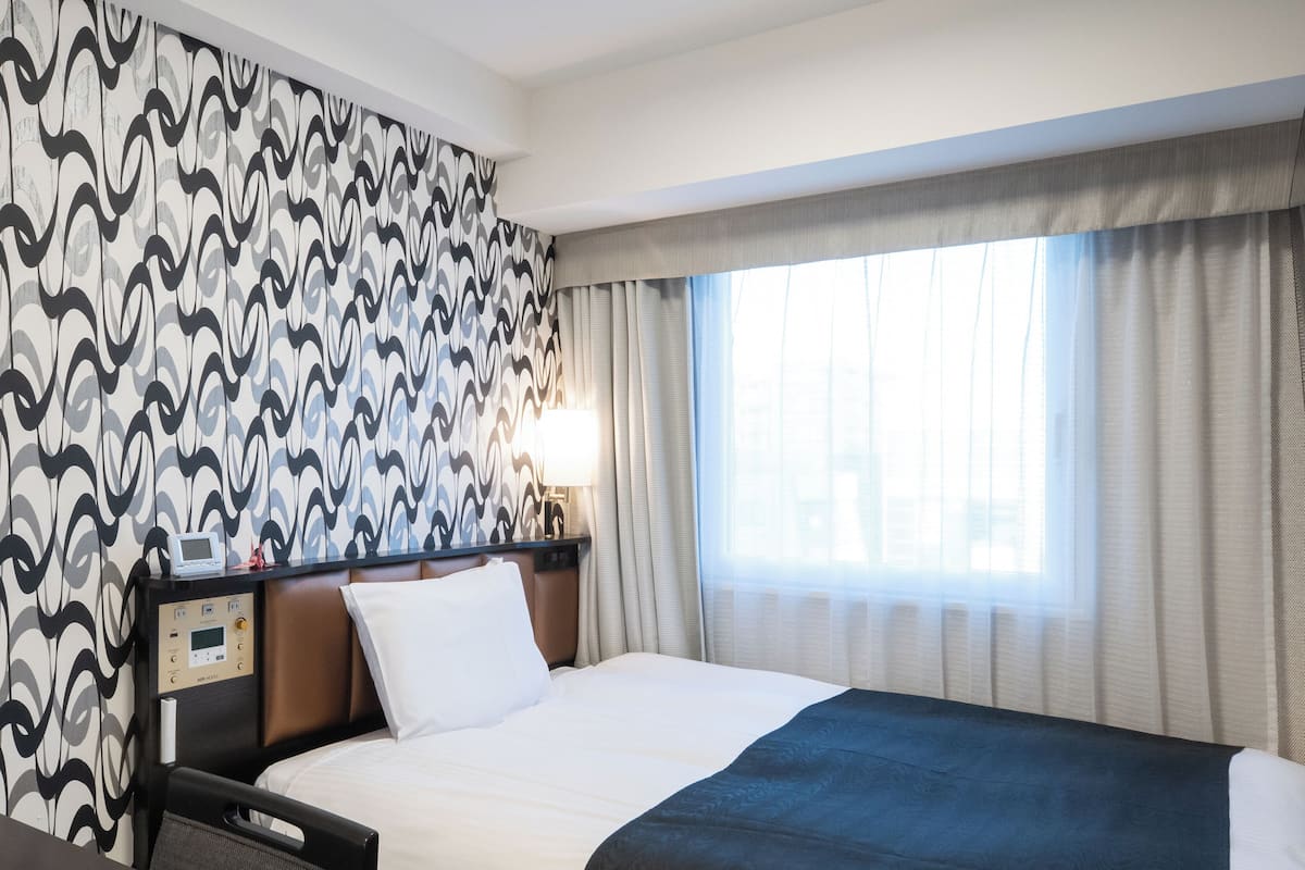 Une chambre d’hôtel avec un lit, un panneau de commande, une table de chevet et une fenêtre avec des rideaux.