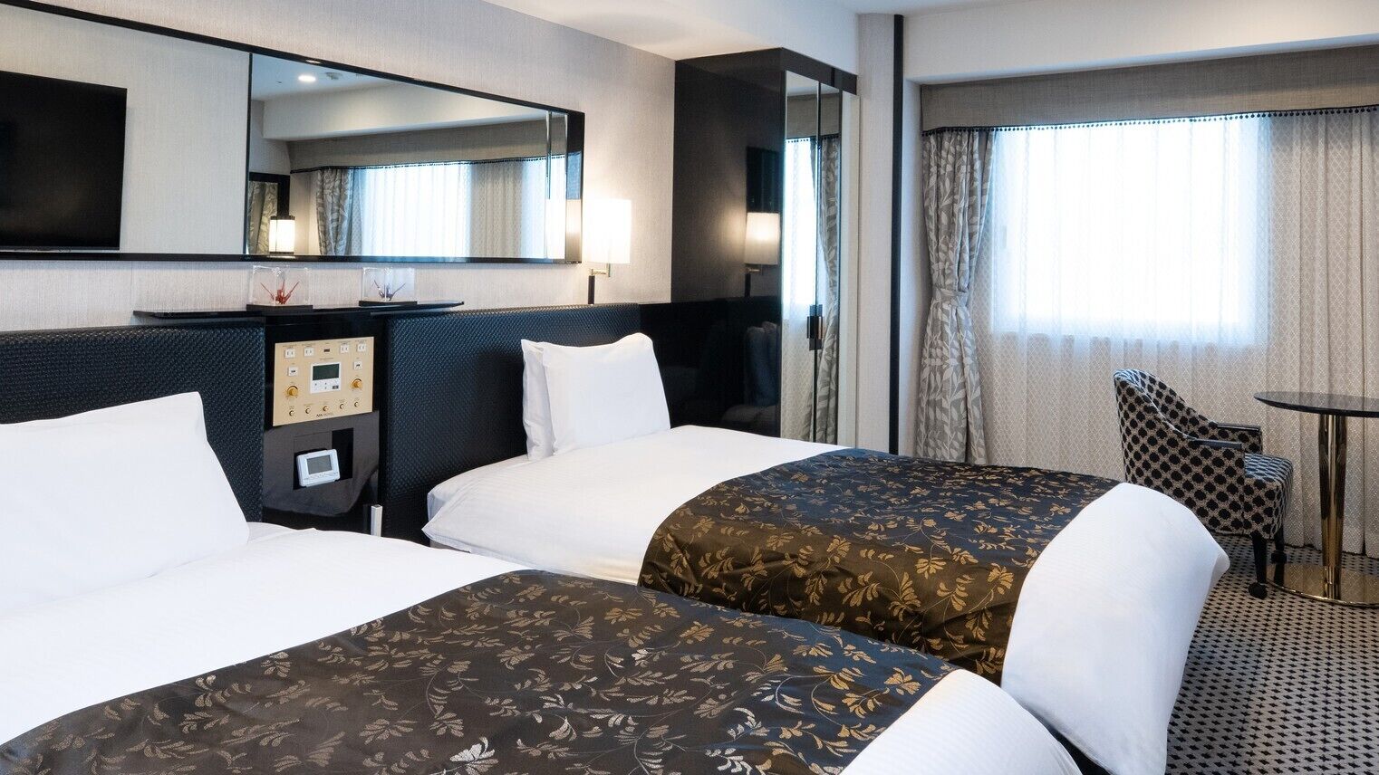 A hotel room with two beds, a desk, a chair, a small table, a mirror, and a window with curtains.