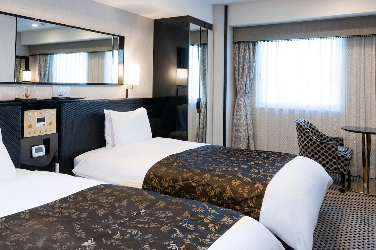 Une chambre d’hôtel avec deux lits, un bureau, une chaise, une petite table, un miroir et une fenêtre avec des rideaux.