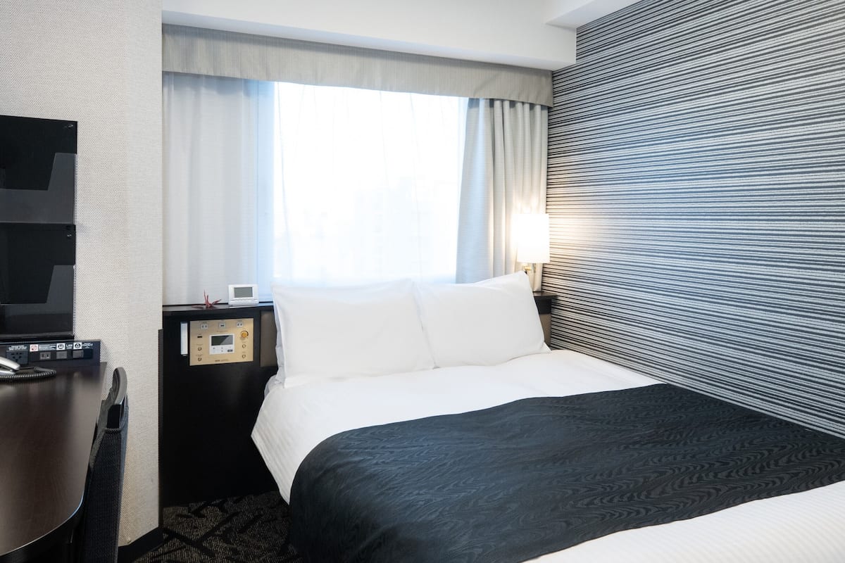 Une chambre d’hôtel comprenant un lit, un bureau, une télévision, une table de chevet avec une lampe et une fenêtre avec des rideaux.