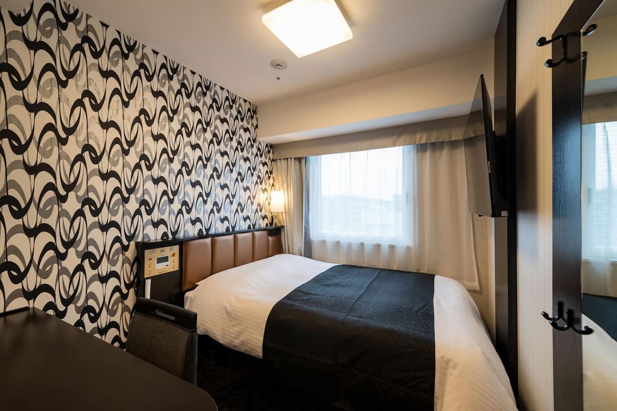 Une chambre d’hôtel avec un mur décoré d’un motif, un lit avec une tête de lit, une table de chevet, une lampe, une fenêtre avec des rideaux et un téléviseur.