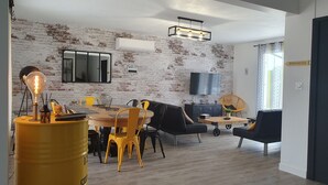 Een moderne woonkamer met een bakstenen muur, een eethoek met gele stoelen, een zwarte bank en een aan de muur bevestigde televisie.