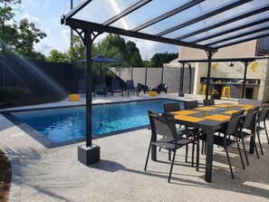 Zona junto a la piscina con un espacio de descanso cubierto, piscina y zona de barbacoa.