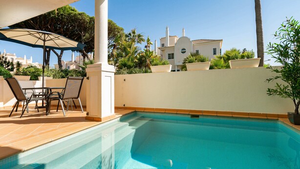 A swimming pool with a sun umbrella, lounge chairs, and a view of a white building with a tiled roof. - Outstanding one bedroom apart with heated private pool in Vale do Lobo (Almancil)