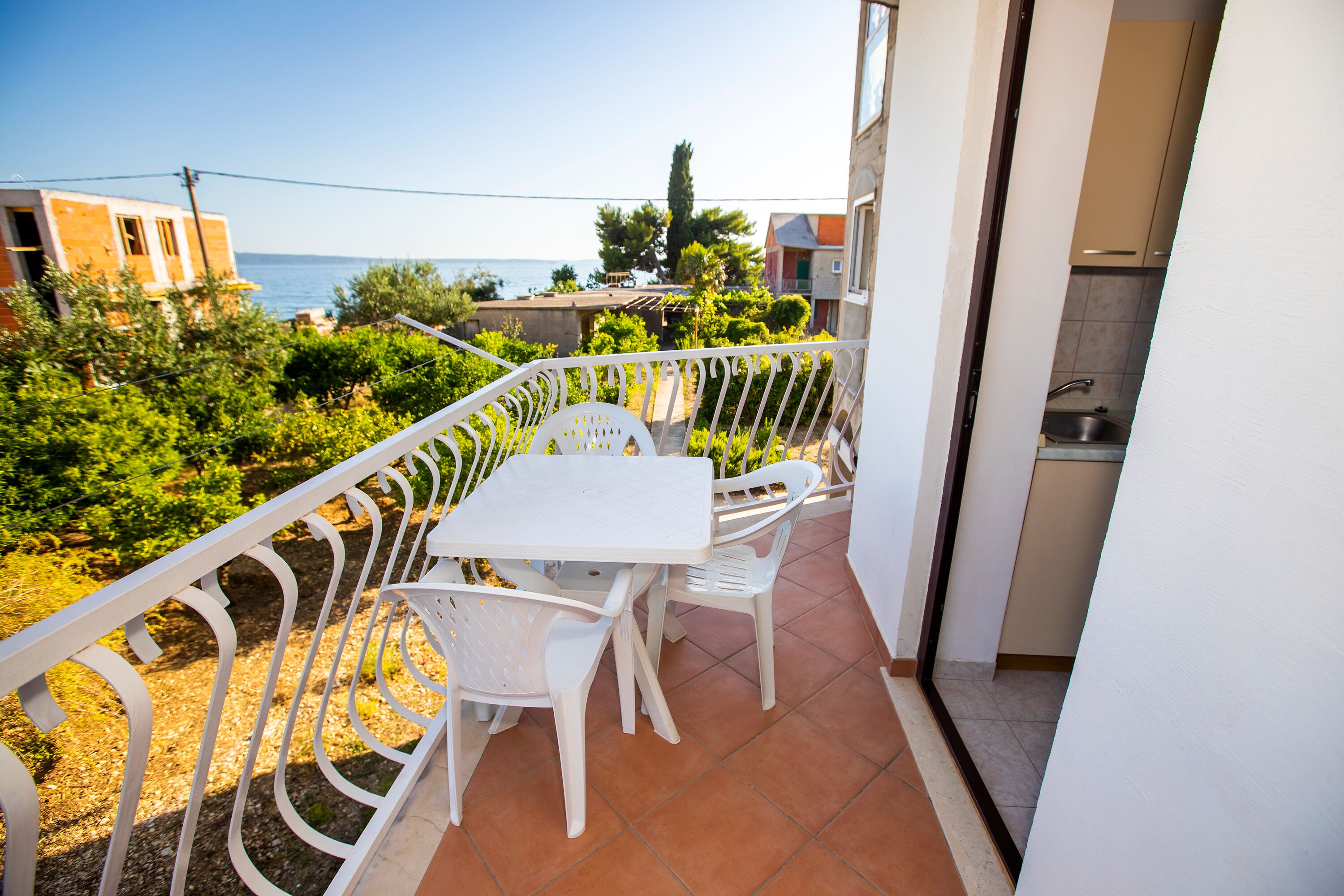 A balcony with a table and chairs, overlooking a garden and the sea.