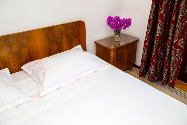 A neatly made bed with white linens and pink floral patterns, a wooden headboard, a small wooden nightstand with a vase of purple flowers, and a red patterned curtain.