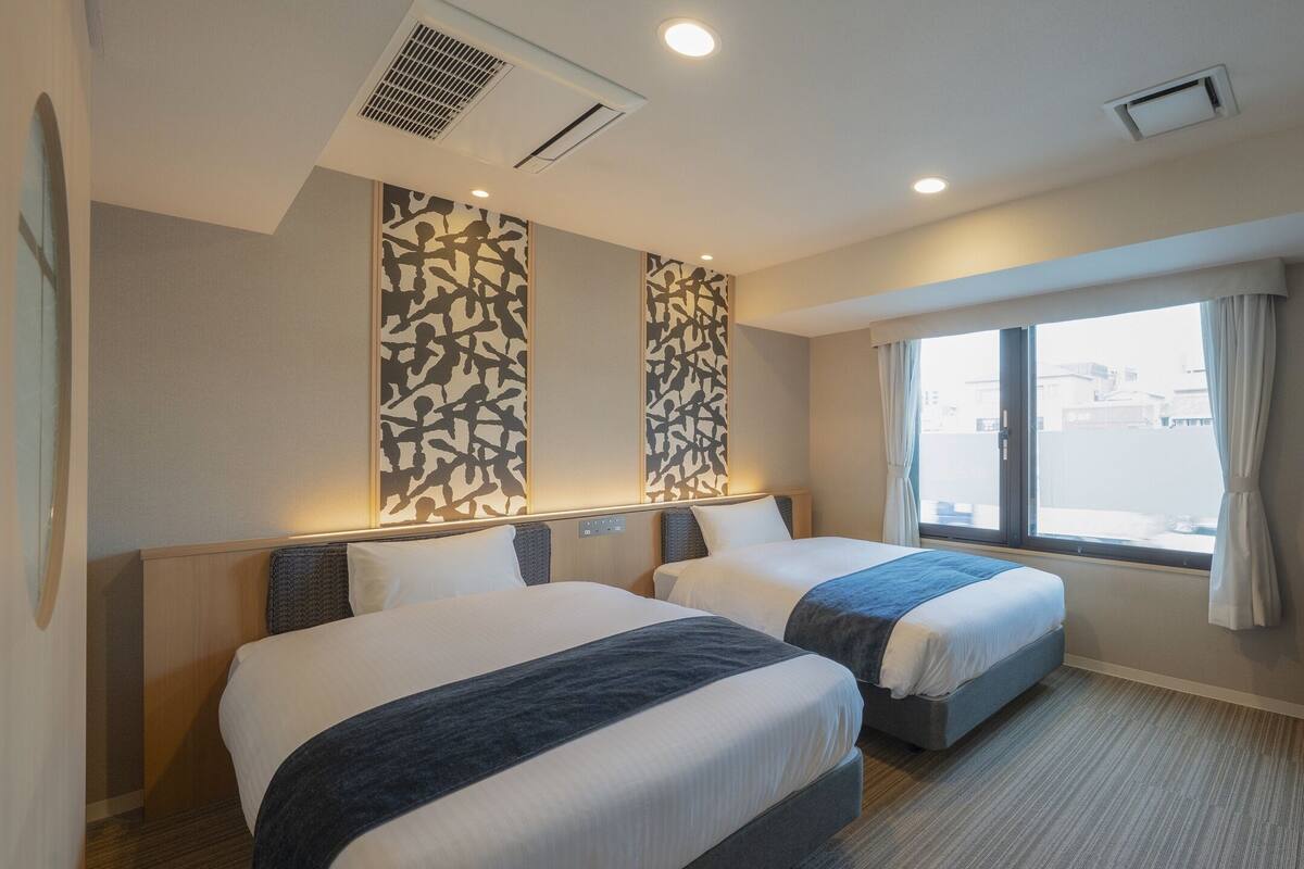 Une chambre d’hôtel avec deux lits, une grande fenêtre avec des rideaux, un téléviseur à écran plat et une armoire.