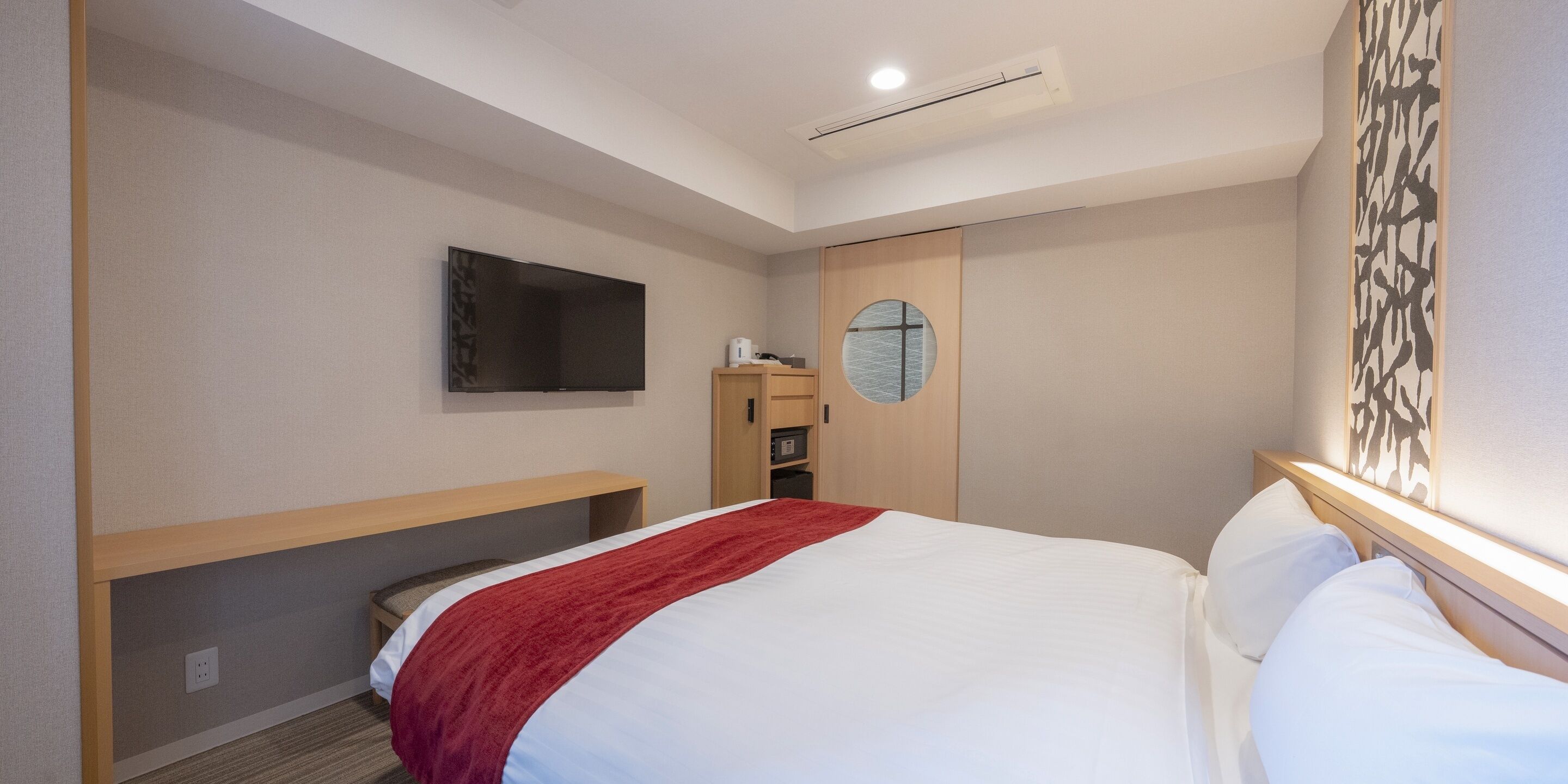 Quarto de hotel com uma cama, TV fixada na parede, um banco de madeira e um armário pequeno.