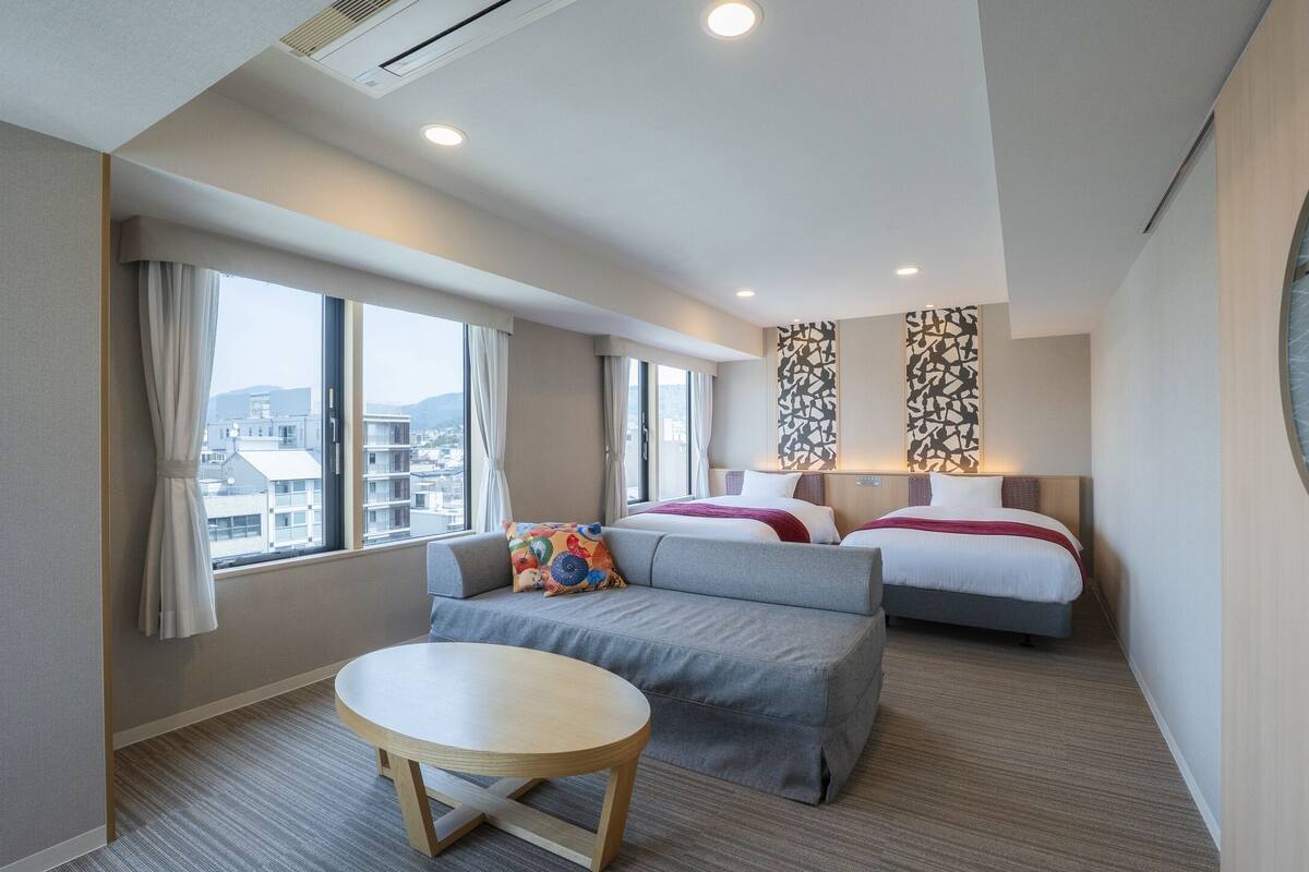 Une chambre d’hôtel avec un canapé, deux lits, une table ronde et de grandes fenêtres donnant sur la ville.
