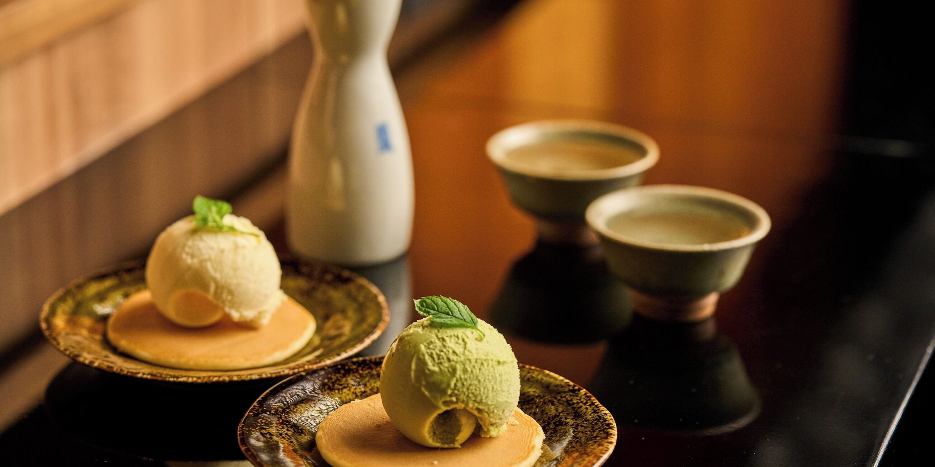 Two desserts on ceramic plates with a vase and two cups in the background.