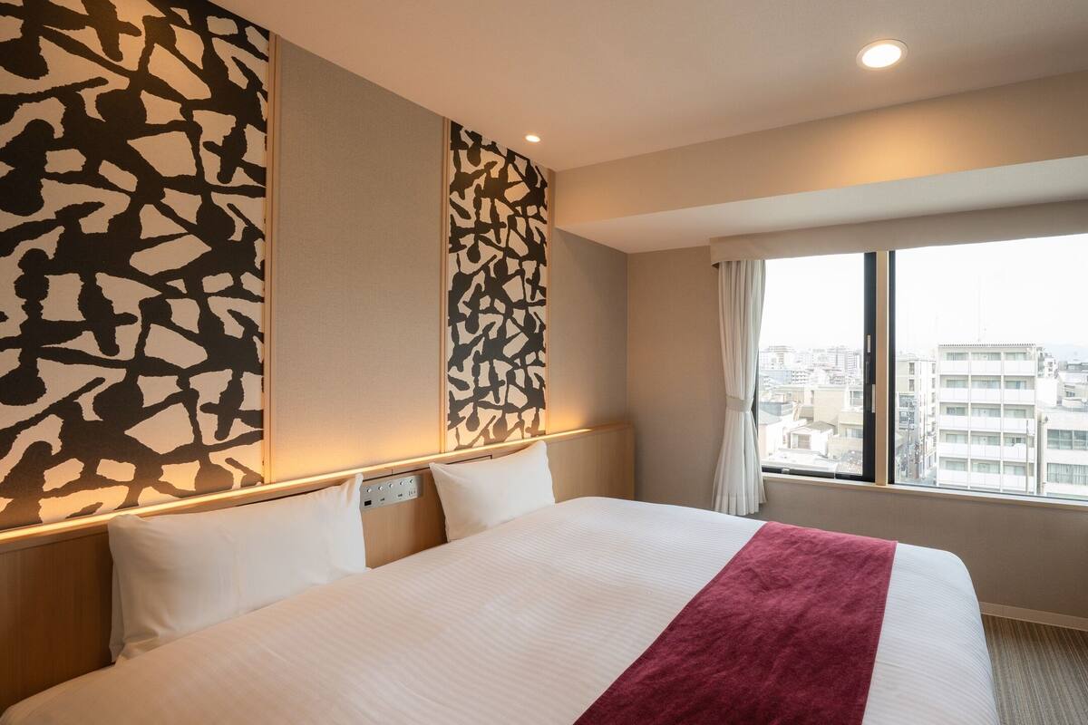 Une chambre d’hôtel avec un grand lit, une vue sur la ville par la fenêtre et un bureau avec un téléphone et des documents.