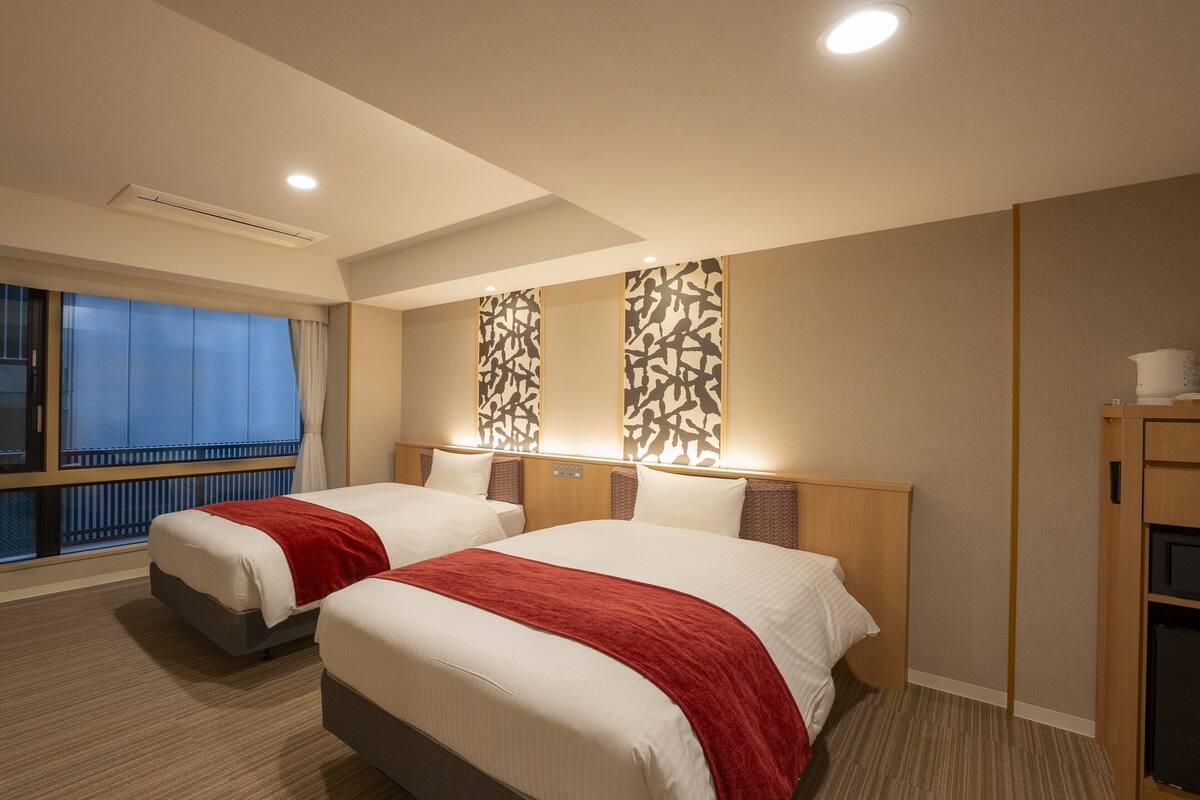 Une chambre d’hôtel avec deux lits, une télévision, une fenêtre avec des rideaux, un climatiseur fixé au mur et une armoire en bois équipée d’un four à micro-ondes.