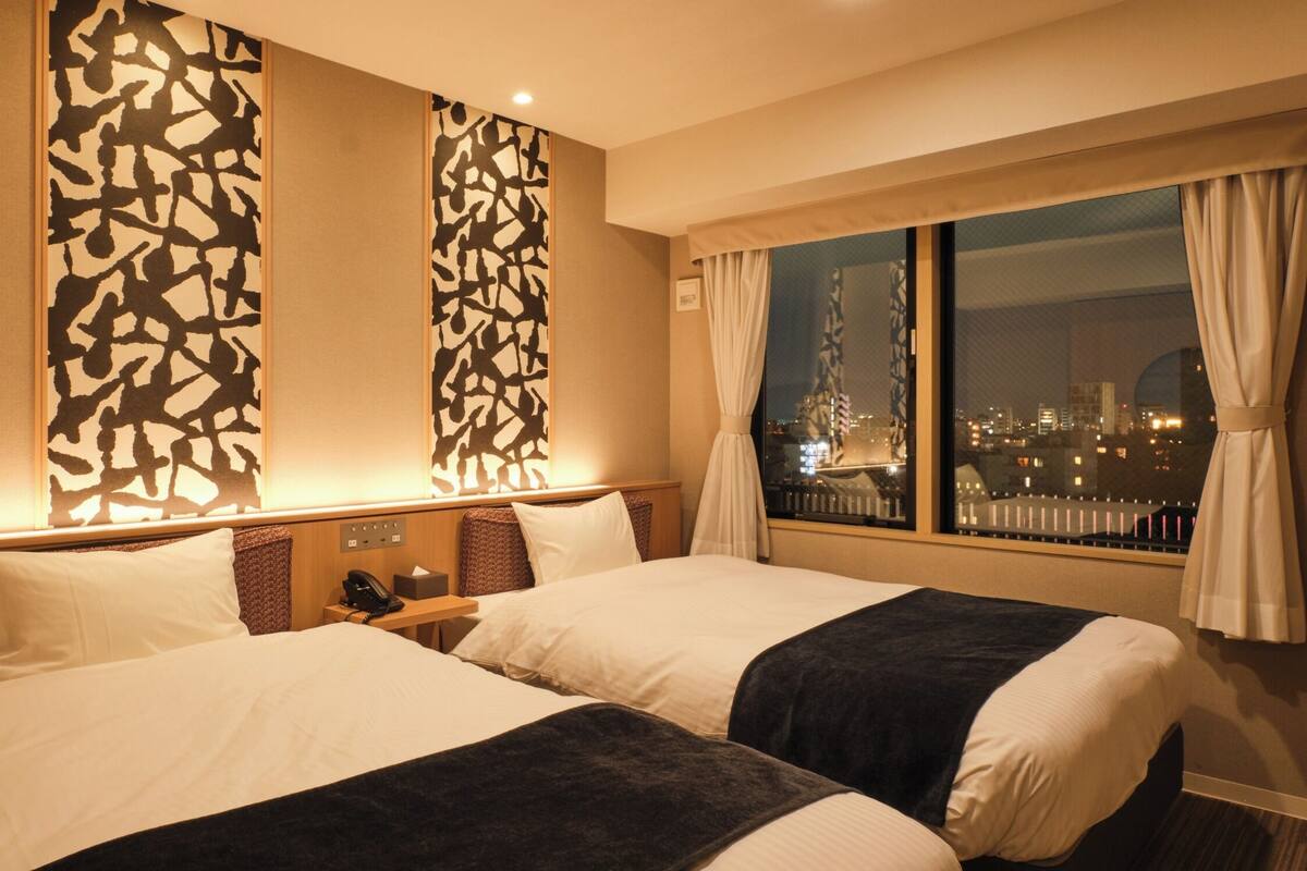 Une chambre d’hôtel avec deux lits, une table de chevet, une fenêtre donnant sur la ville et un mur décoré d’un motif.
