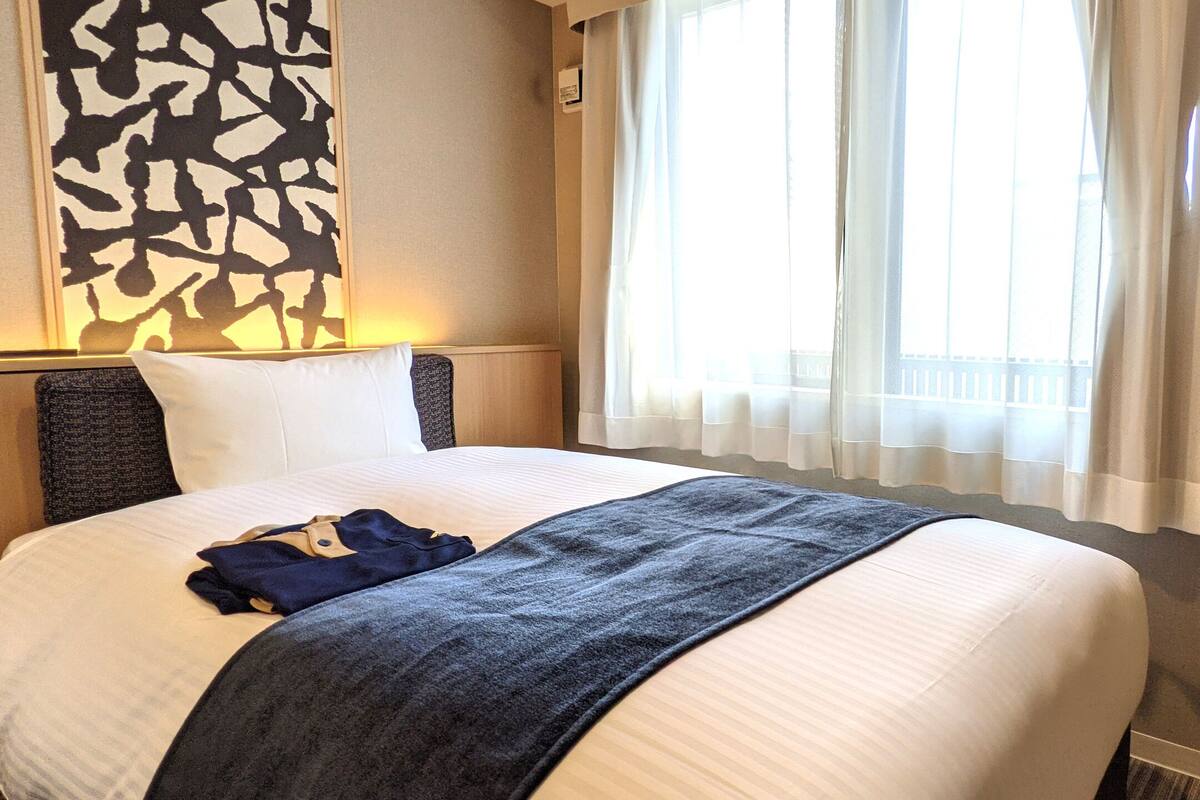 Un lit bien fait, avec une couette blanche et une couverture bleu foncé, un oreiller et un vêtement plié posé sur le lit.