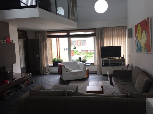 A modern living room with a white sofa, a beige sofa, a coffee table, and a TV on a stand. There is a large window with curtains, a balcony, and a painting on the wall.