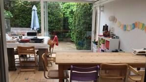 A covered patio area with a wooden table and chairs, a bookshelf, and a view of the garden. - Grote Familiewoning Heel Dicht bij het Strand (Noordwijk Zh)