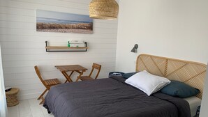 A bedroom with a bed, a small table, two chairs, a hanging light fixture, a wall-mounted shelf with a printer, and a framed picture of a beach.