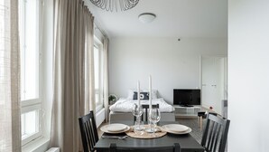 Ein modernes, helles Zimmer mit Esstisch für Zwei, einem Bett mit Fernseher und einem großen Fenster mit Vorhängen.