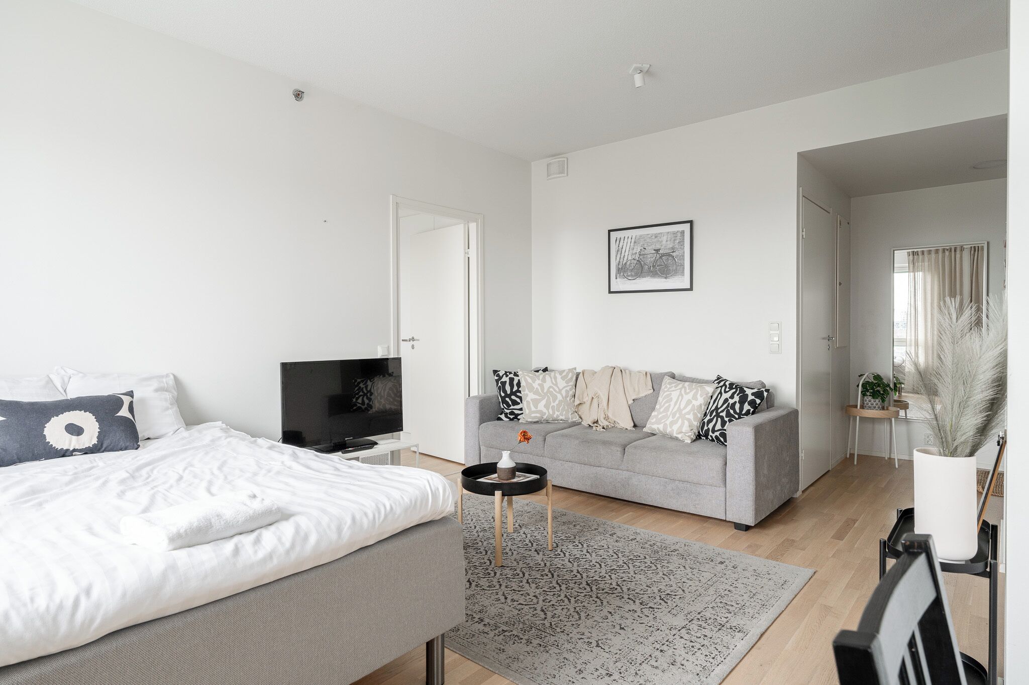 Ein modernes, helles Schlafzimmer mit einem Bett, einem Fernseher und einem grauen Sofa.
