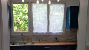 A kitchen with a tiled floor, a window with curtains, a sink, and cabinets. - Maison de Vacance Tout Confort Avec Piscine (saint benoit du sault)